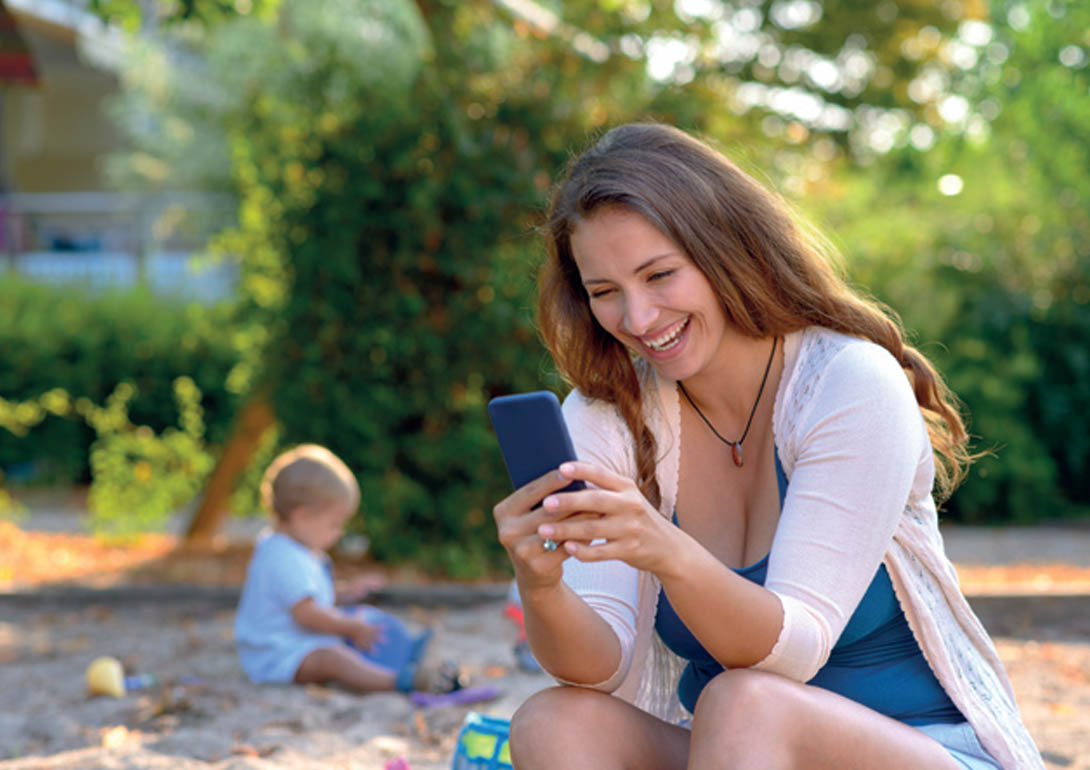 Eine Mutter lächelt ihr Handy an, während ein kleines Kind allein im Hintergrund im Sandkasten sitzt.